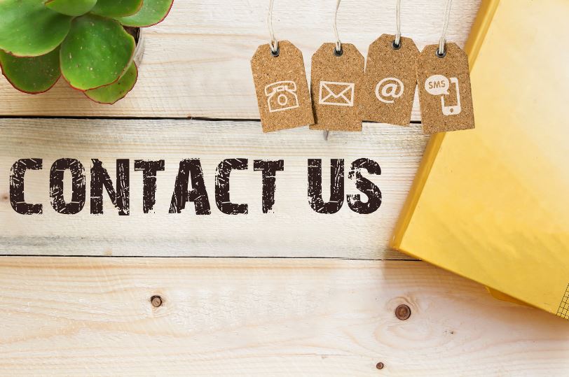 Wooden crate with "Contact Us" stamped in bold lettering, envelopes, and a plant. Four cork luggage labels with icons of phone, envelope, @, and SMS. Learn English by contacting us today!